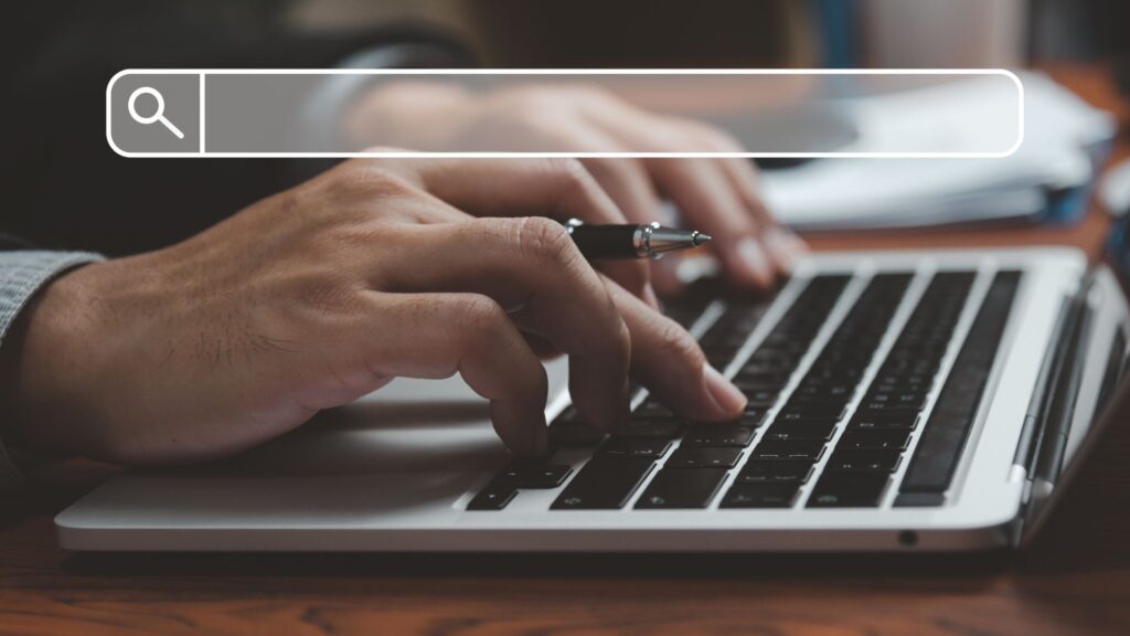 Close-up das mãos de uma pessoa digitando em um teclado de laptop com uma caneta, simbolizando a pesquisa online e o trabalho em SEO.